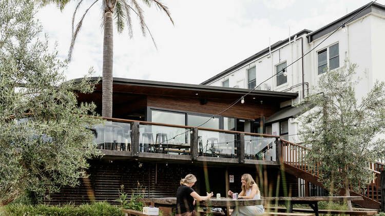 Two people sit at an outdoor picnic table on the wooden deck of a modern building surrounded by trees and a palm tree — SeaChange Parks Milton in Milton, NSW