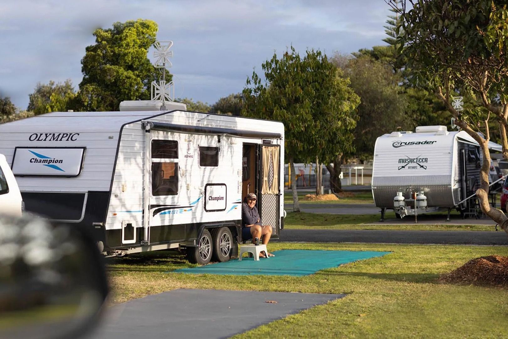 Two RVs parked at a campsite; a person sits outside one. Green grass, trees, and blue sky. — SeaChange Parks Milton in Lake Conjola, NSW