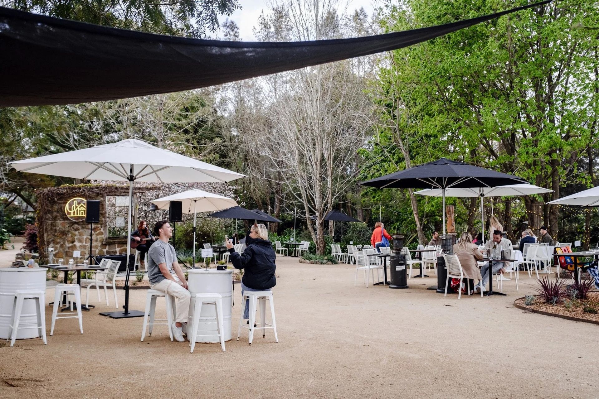 Patrons sit at outdoor tables with umbrellas in a gravel garden café, with a musician performing in the background — SeaChange Parks Milton in Milton, NSW