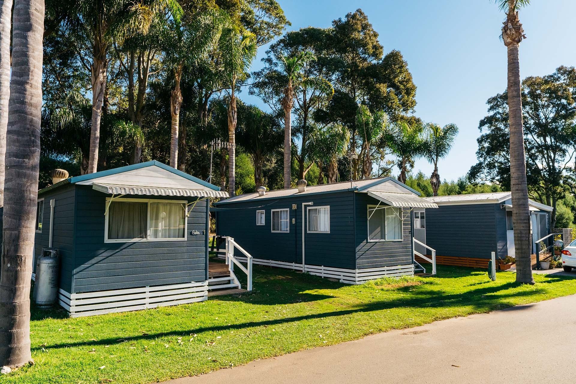 Row of small grey cabins with white trim beside a sunlit road — SeaChange Parks Milton in Milton Park, NSW