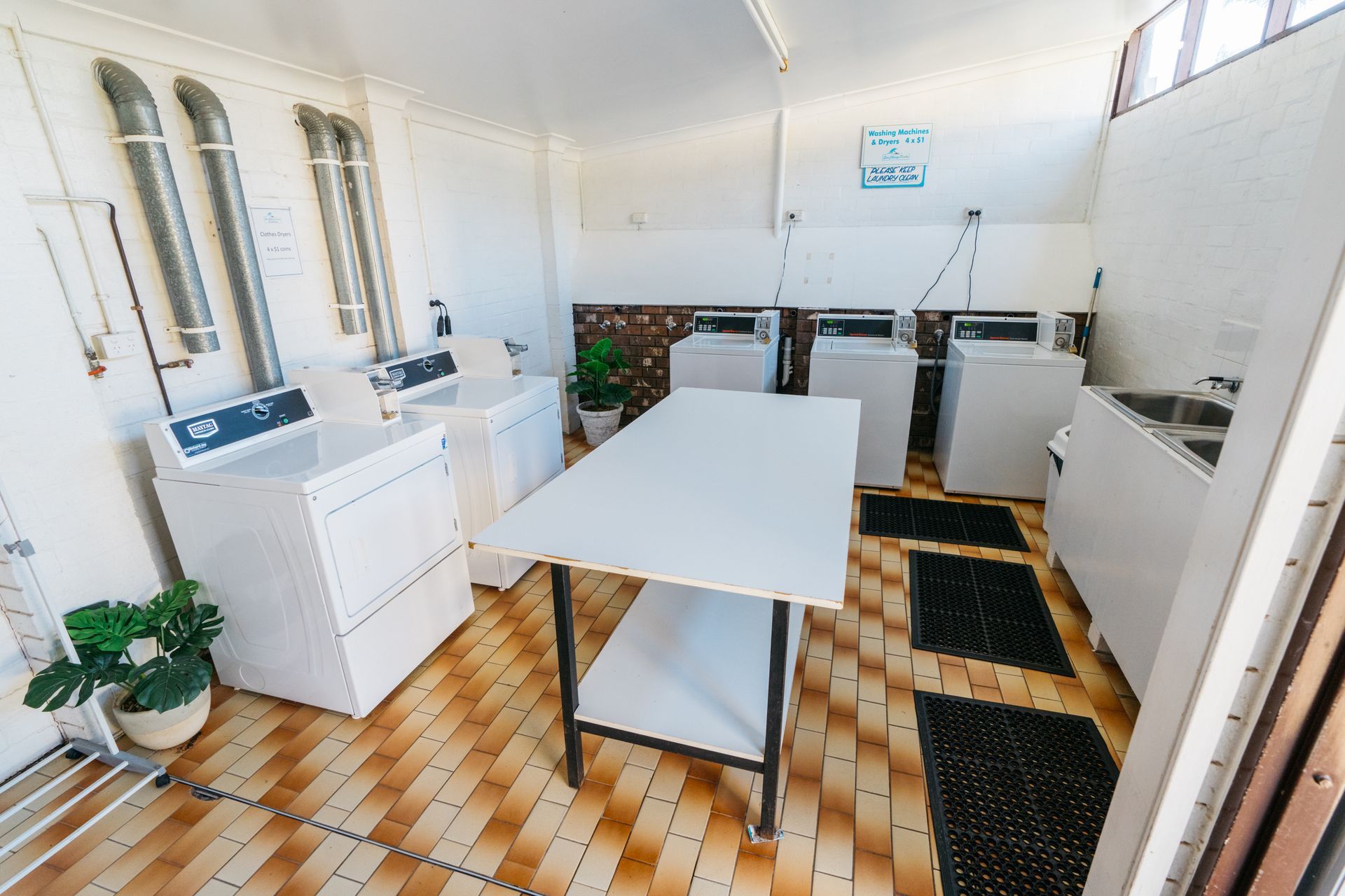 A bright laundry room with white machines, a center folding table, and tan tiled floors  — SeaChange Parks Milton in Milton, NSW