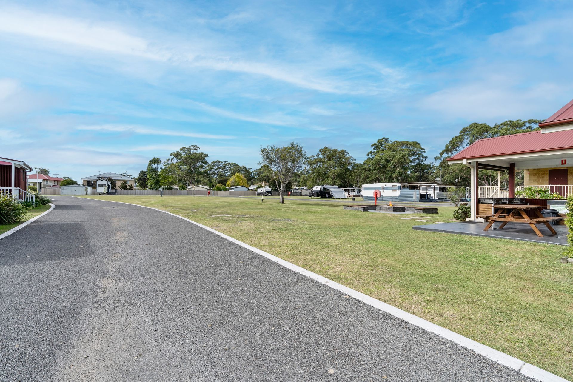 A gravel road leads past a grassy lawn toward cabins and RVs under a bright blue sky at a holiday park — SeaChange Parks Milton in Lake Conjola, NSW
