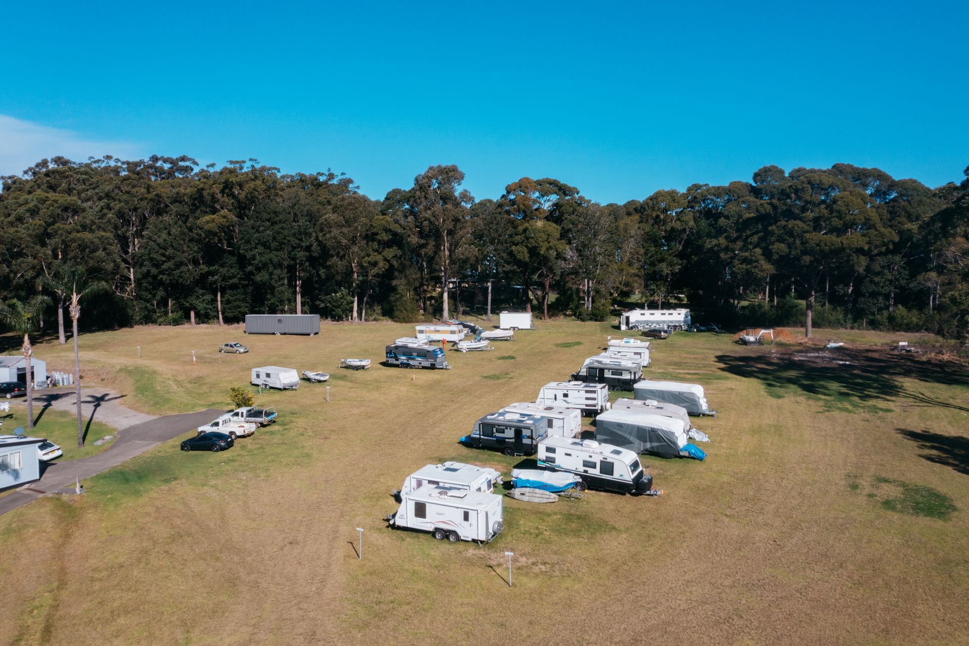 An aerial view of a grassy campground filled with parked white travel trailers and RVs bordered by dense forest trees — SeaChange Parks Milton in Milton, NSW