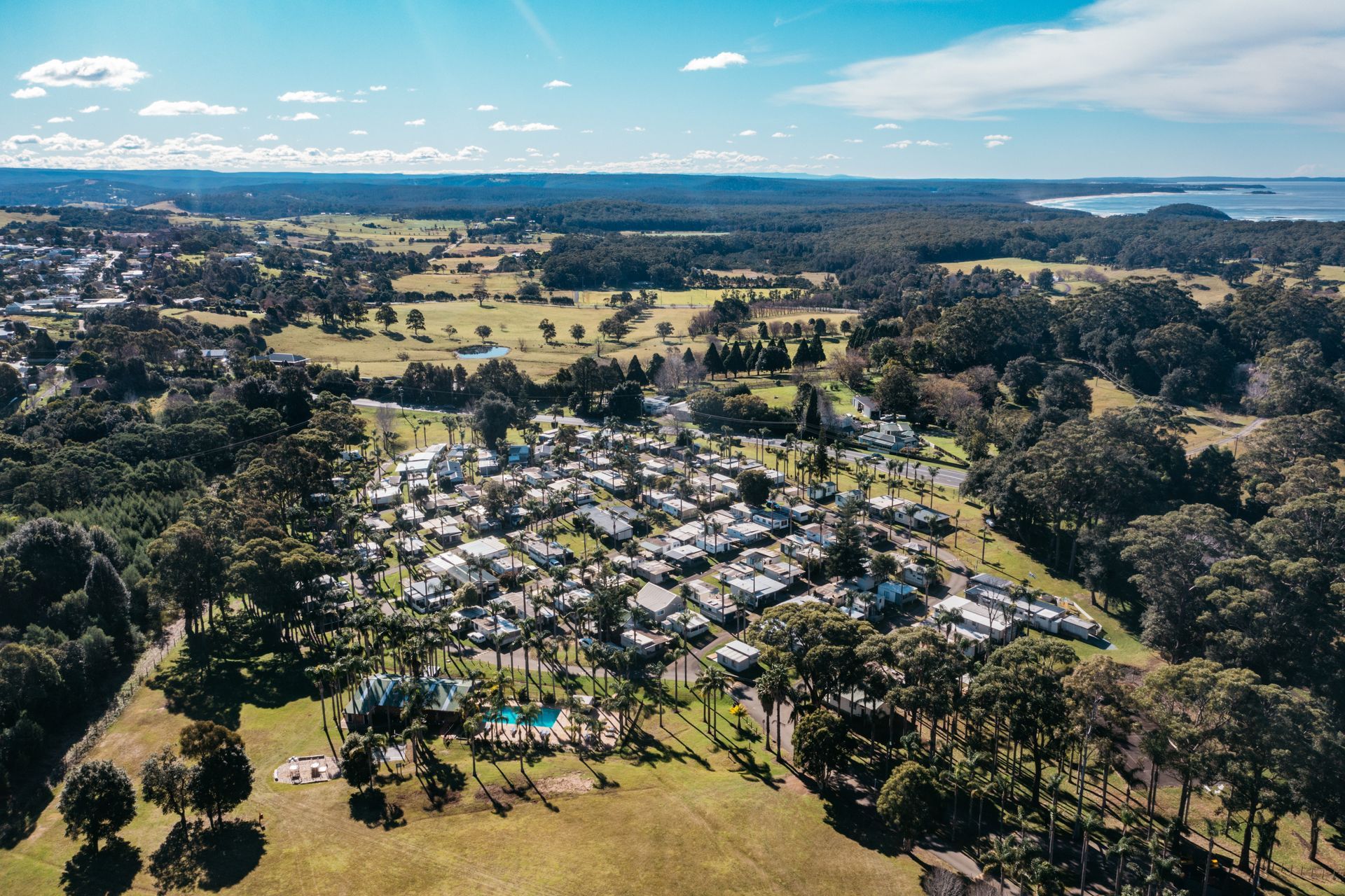 An aerial view shows a dense cluster of small homes and cabins set among trees with a swimming pool and coastal backdrop  — SeaChange Parks Milton in Milton, NSW
