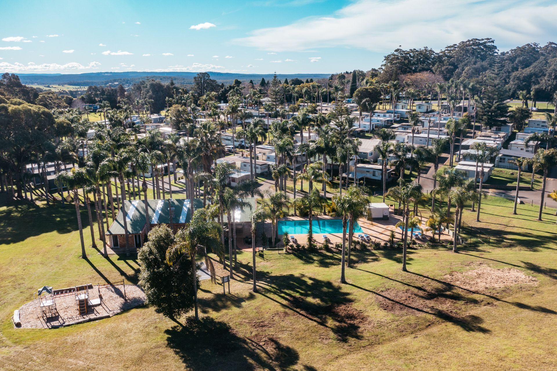 An aerial view of a resort with a swimming pool surrounded by palm trees, lawn areas, and rows of cabins in the distance  — SeaChange Parks Milton in Milton, NSW