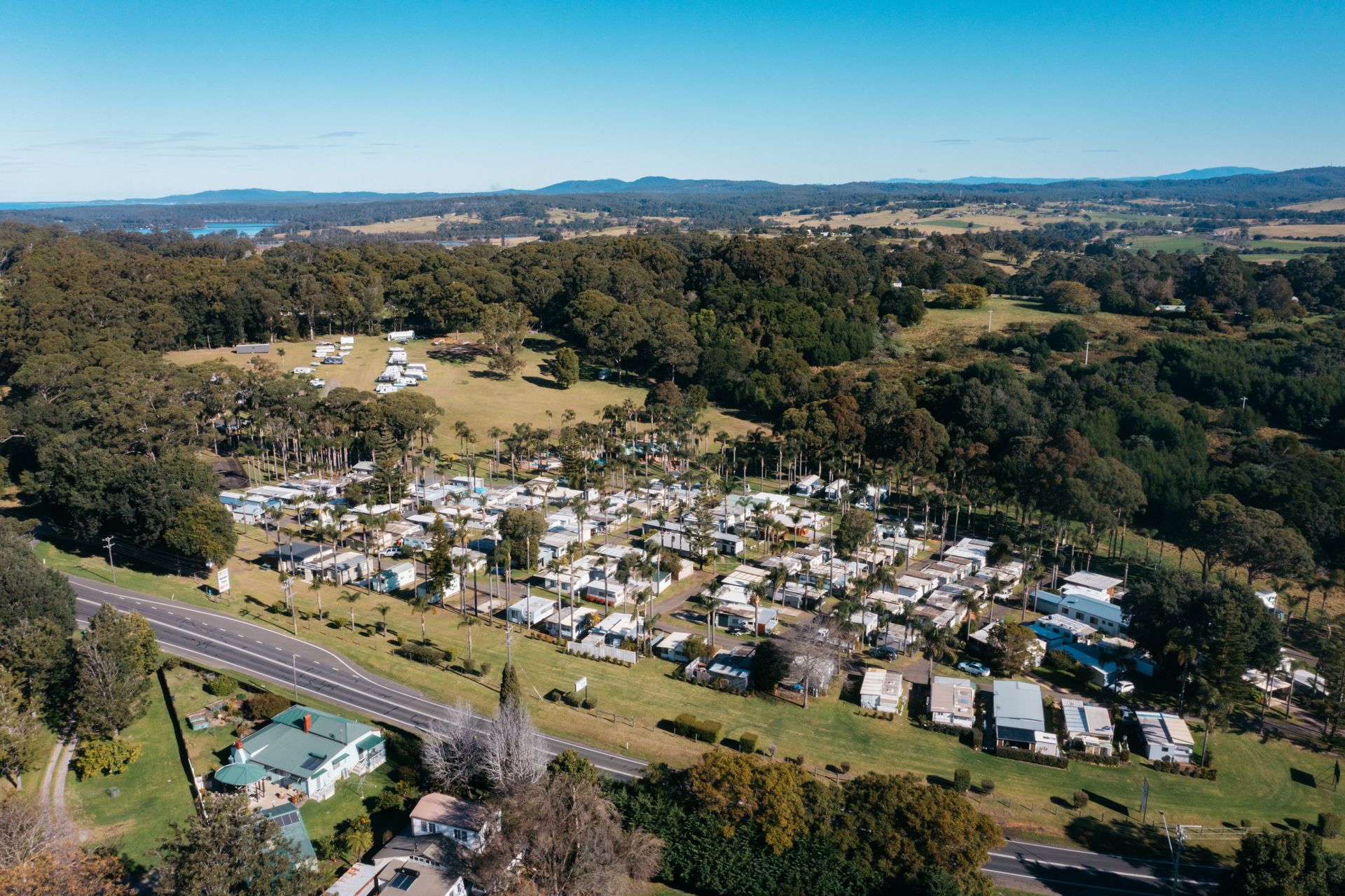 Aerial view of a densely packed caravan park set among trees and rolling green hills under a bright blue sky  — SeaChange Parks Milton in Milton, NSW