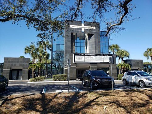 Modern building with a cross-shaped design, glass windows, and a sign. Palm trees and parked cars are in front.