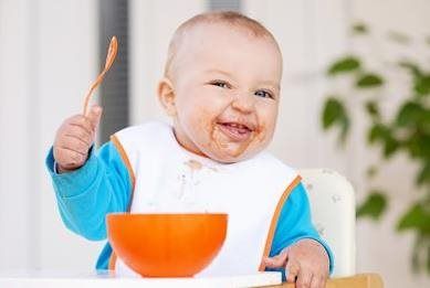 Smiling baby covered in food, holding spoon, sitting at a table with bowl.