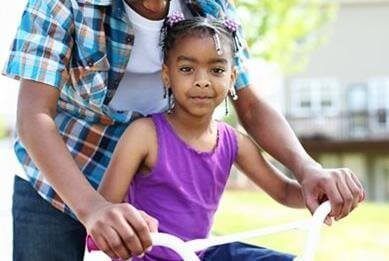 Person helping a child ride a bicycle outdoors; girl smiles, wearing purple.