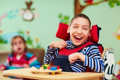 Boy in a wheelchair smiles broadly at a desk with colorful toy blocks. Another child in the background.