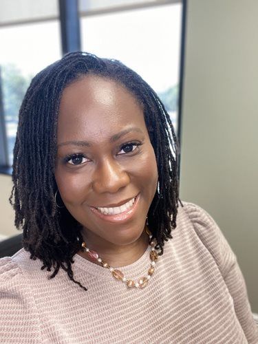Woman with locs smiles at the camera, wearing a light pink sweater and necklace.