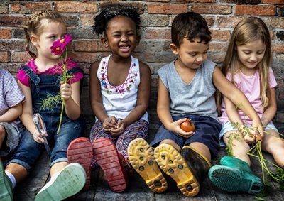 Children sitting together by brick wall, holding flowers and vegetables, smiling.