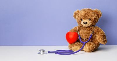 Teddy bear with a stethoscope and red heart against a purple background, symbolizing healthcare.