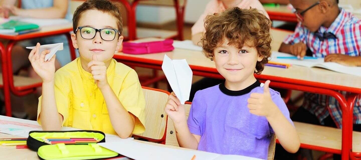 Two children in a classroom hold paper airplanes, smiling. One gives a thumbs up.
