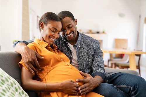 Couple smiling, holding pregnant belly on a couch indoors.