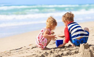 Two children playing with sand and a blue bucket on a beach, waves in the background.