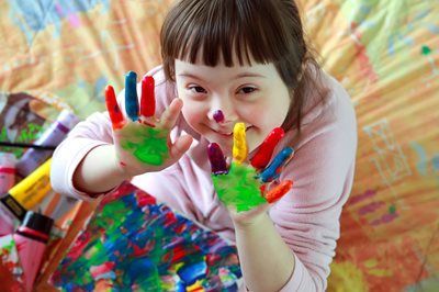 Child with Down syndrome smiles, holding up paint-covered hands. Colorful artwork in background.