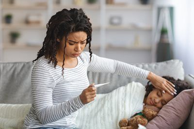 Woman taking a child's temperature with a thermometer; they are on a couch indoors.