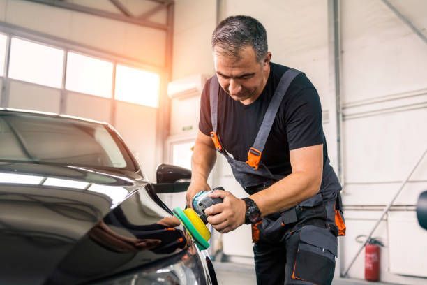 Car service worker polishes the surface of a black car with a polisher inside an auto repair shop.