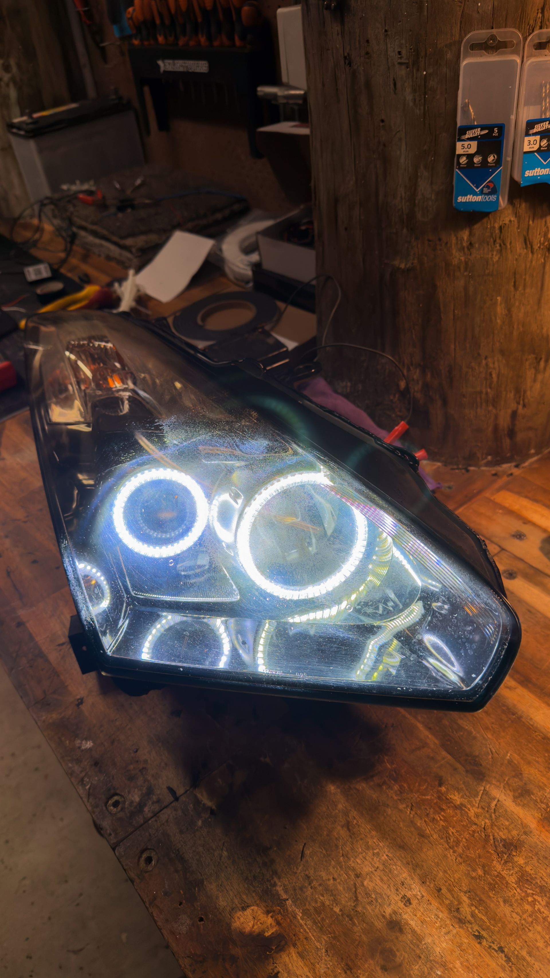 A custom automotive headlight assembly with illuminated white LED halo rings sits on a wooden workbench.