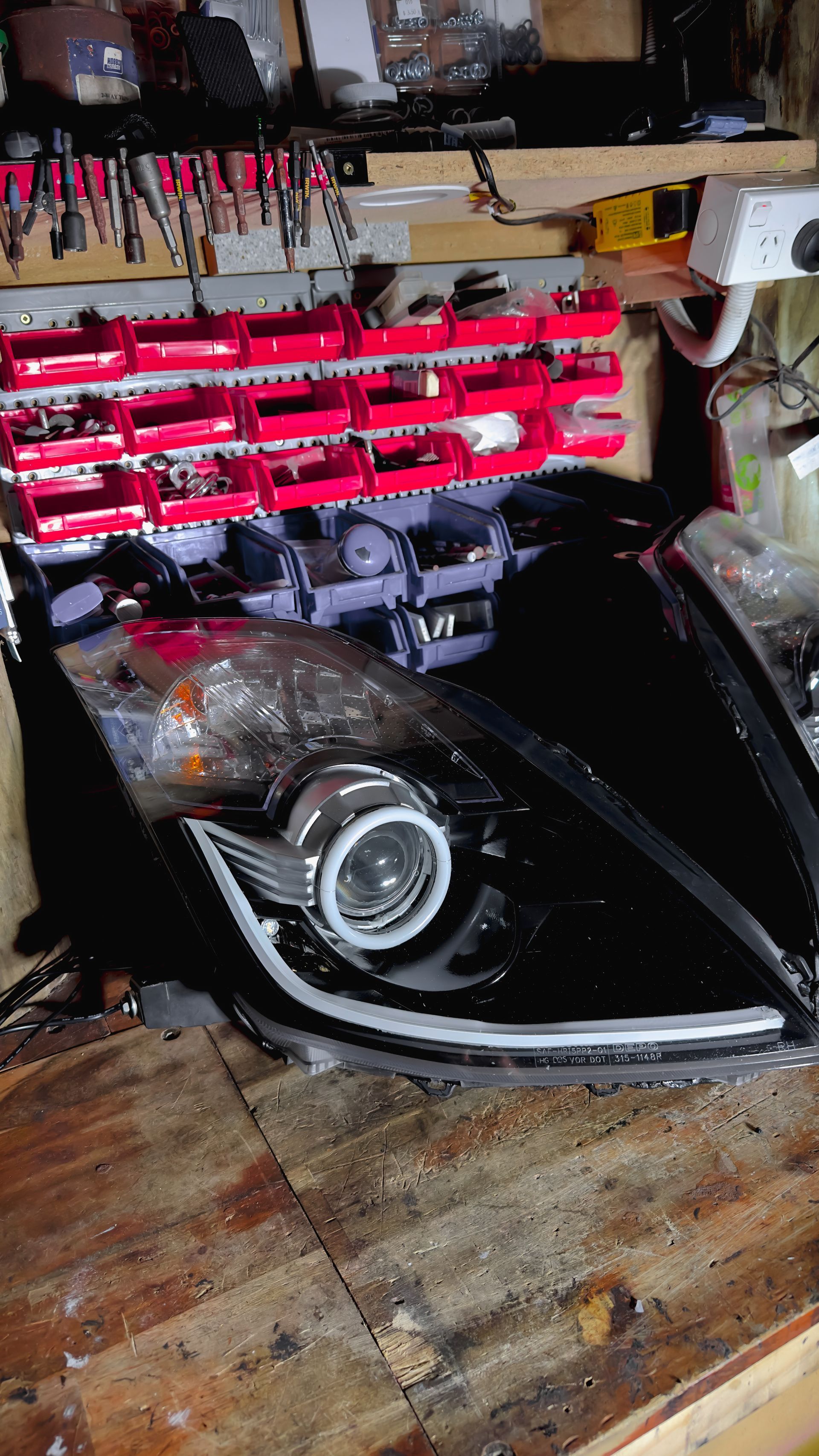 Black car headlight with a white halo and LED strip on a wooden surface in a workshop.