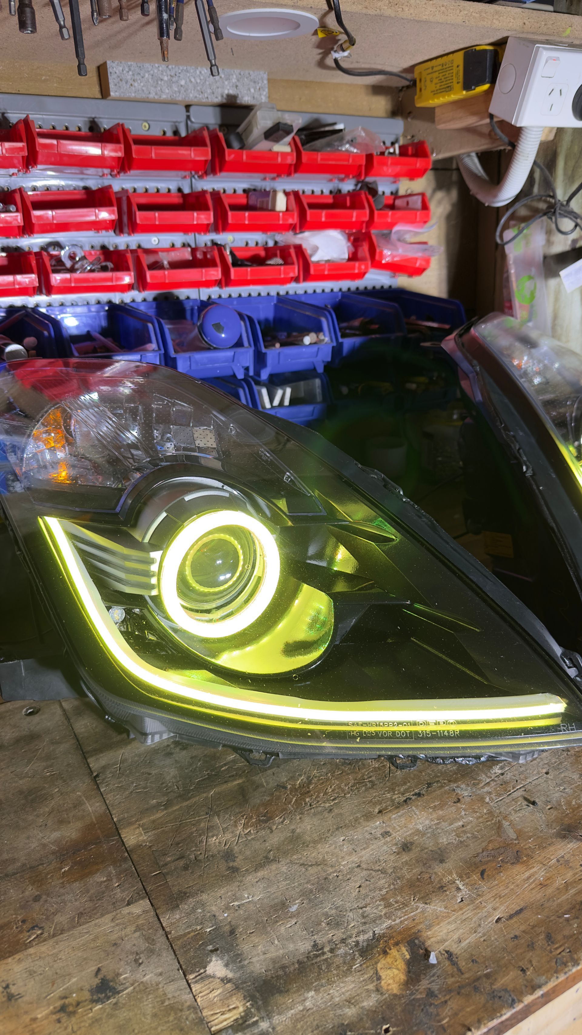 Black car headlight with a white halo and LED strip on a wooden surface in a workshop.