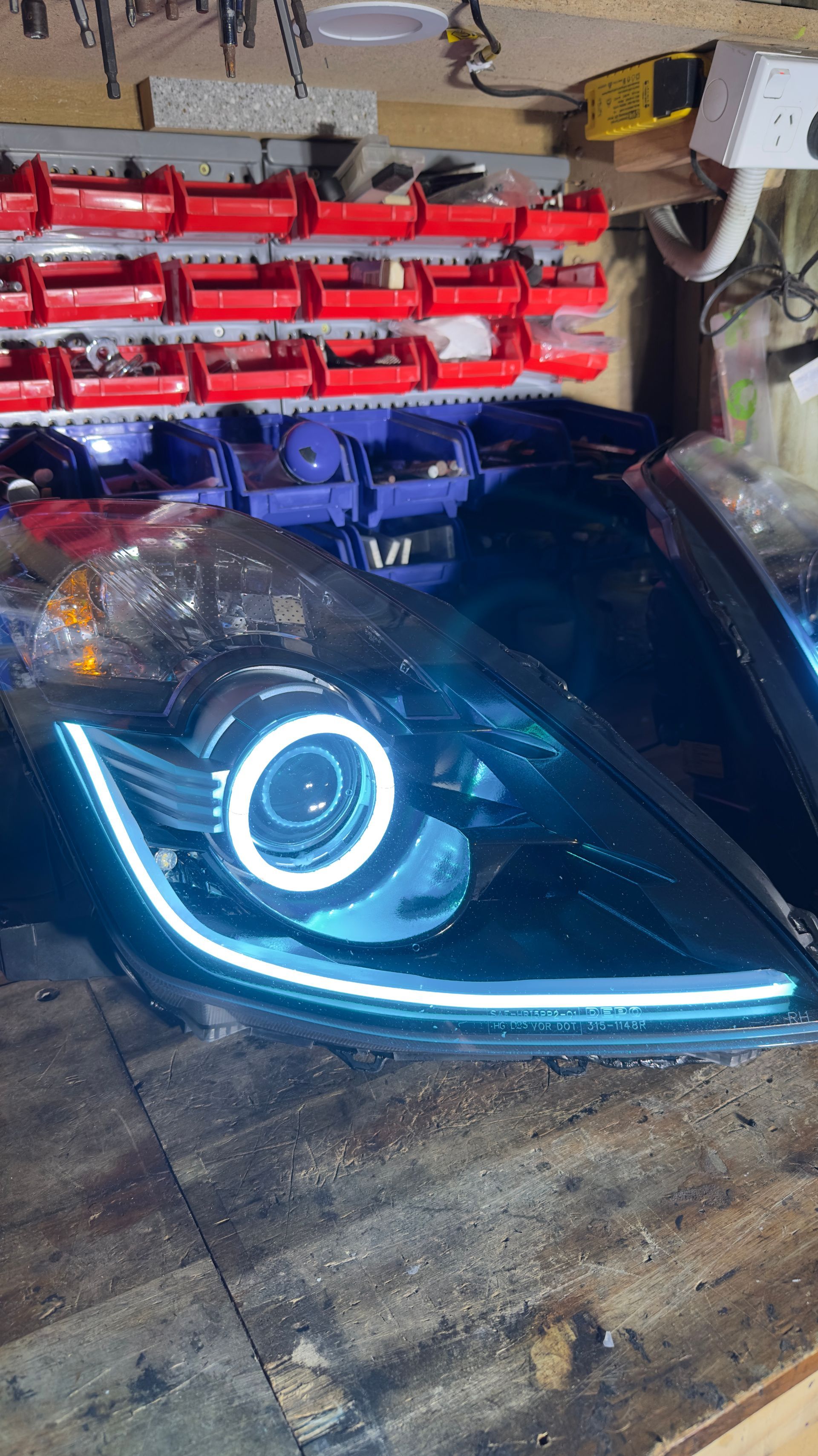 Black car headlight with a white halo and LED strip on a wooden surface in a workshop.