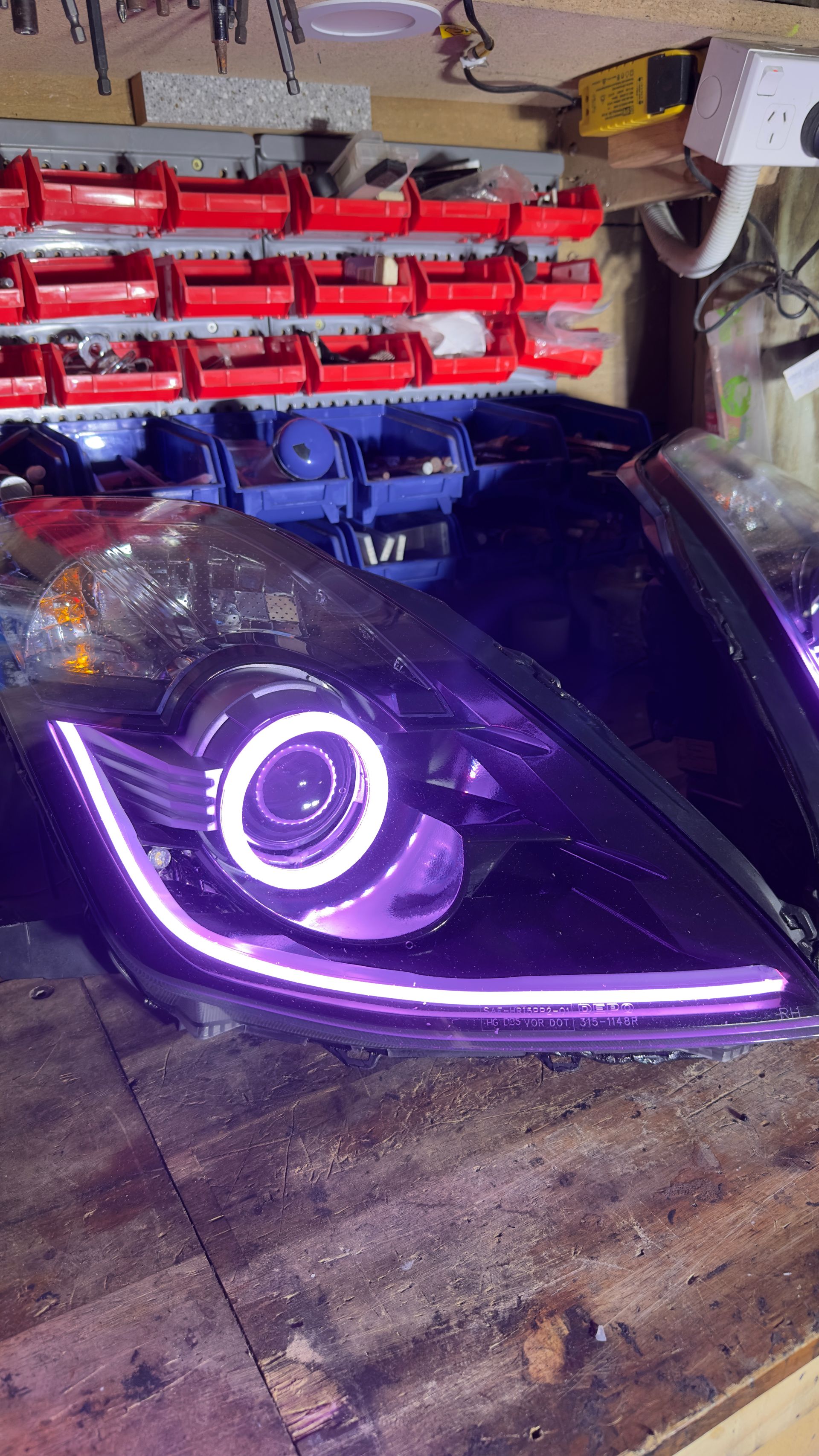 Black car headlight with a white halo and LED strip on a wooden surface in a workshop.