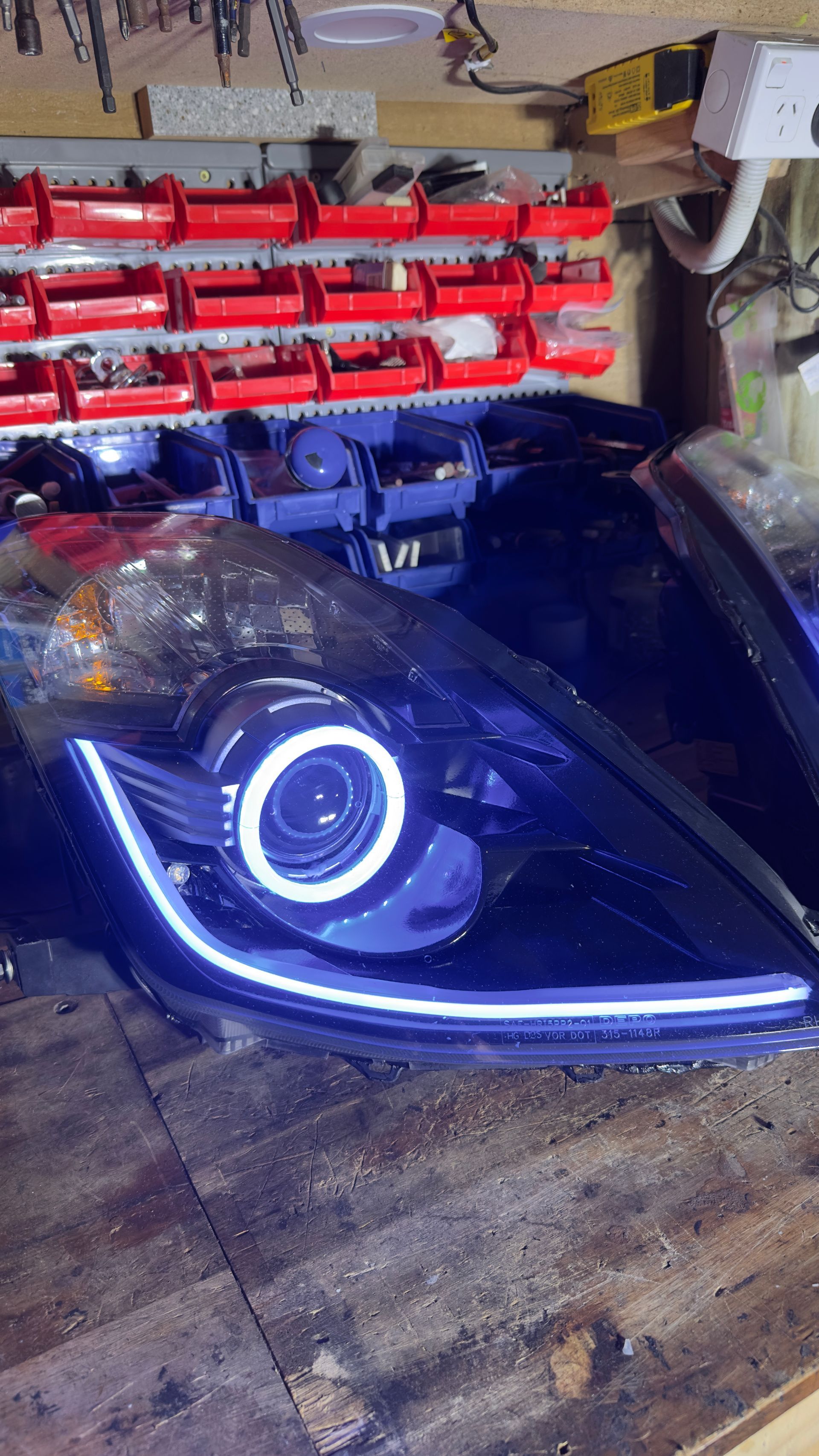 Black car headlight with a white halo and LED strip on a wooden surface in a workshop.