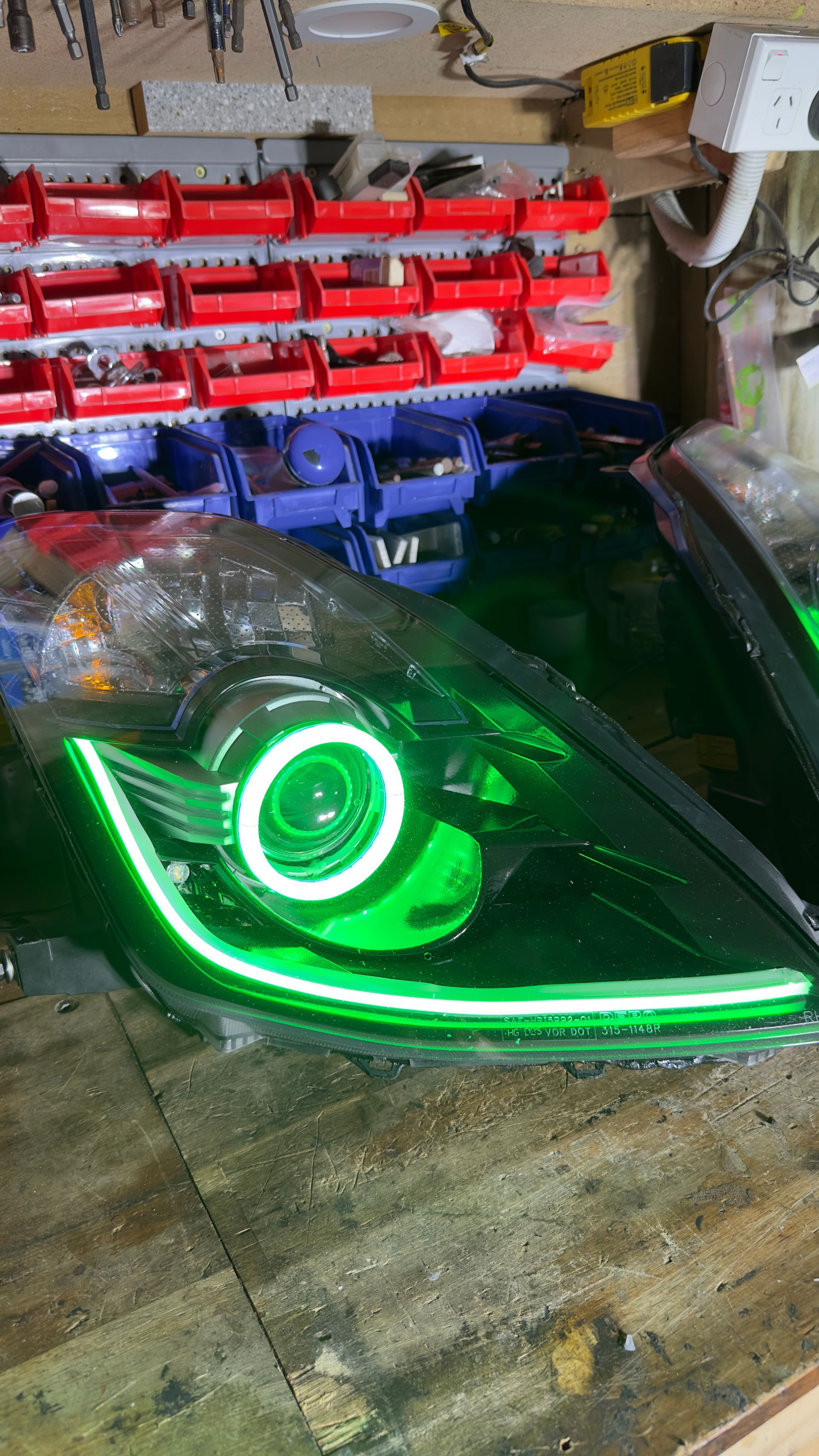 Black car headlight with a white halo and LED strip on a wooden surface in a workshop.