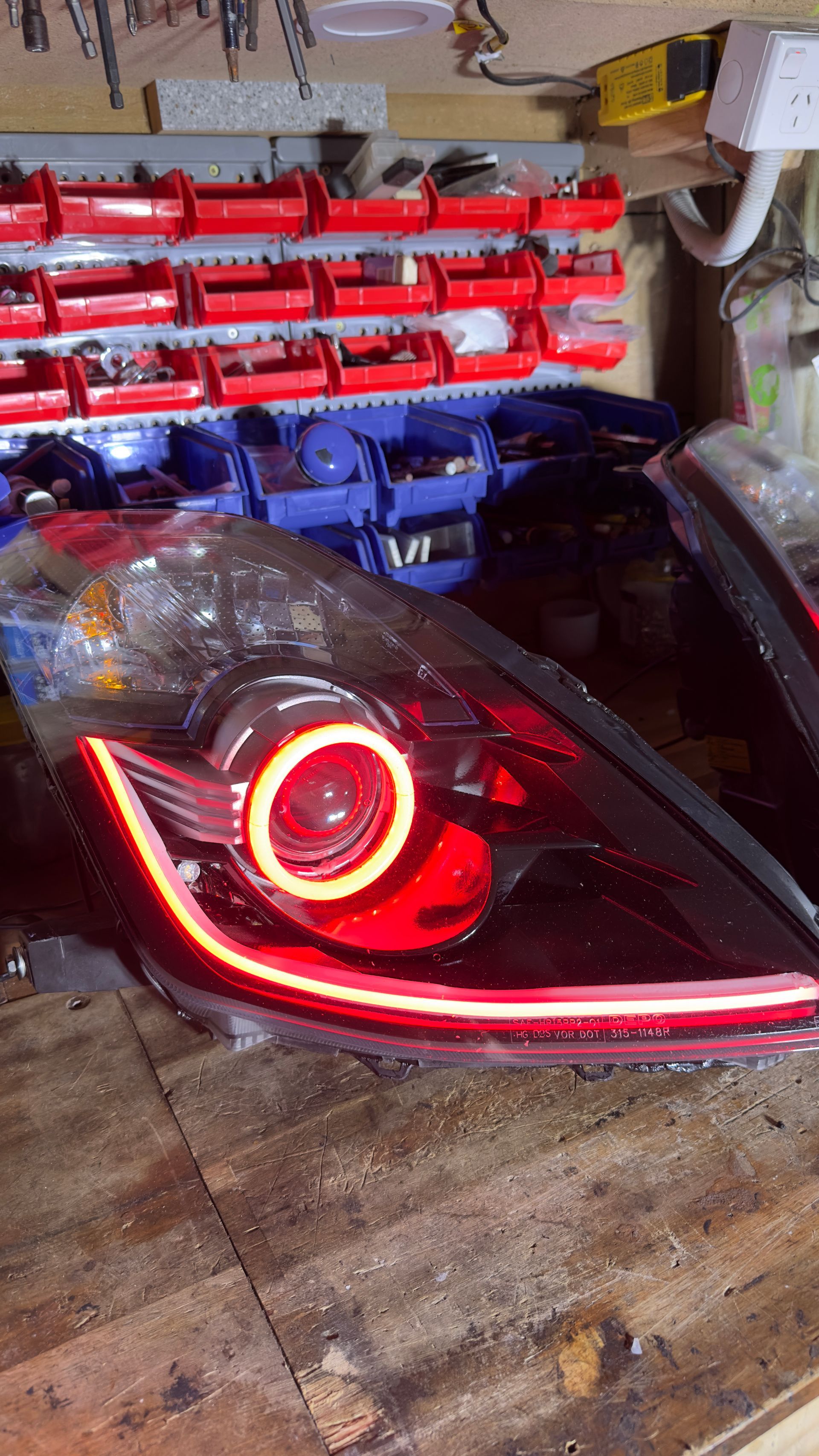 Black car headlight with a white halo and LED strip on a wooden surface in a workshop.