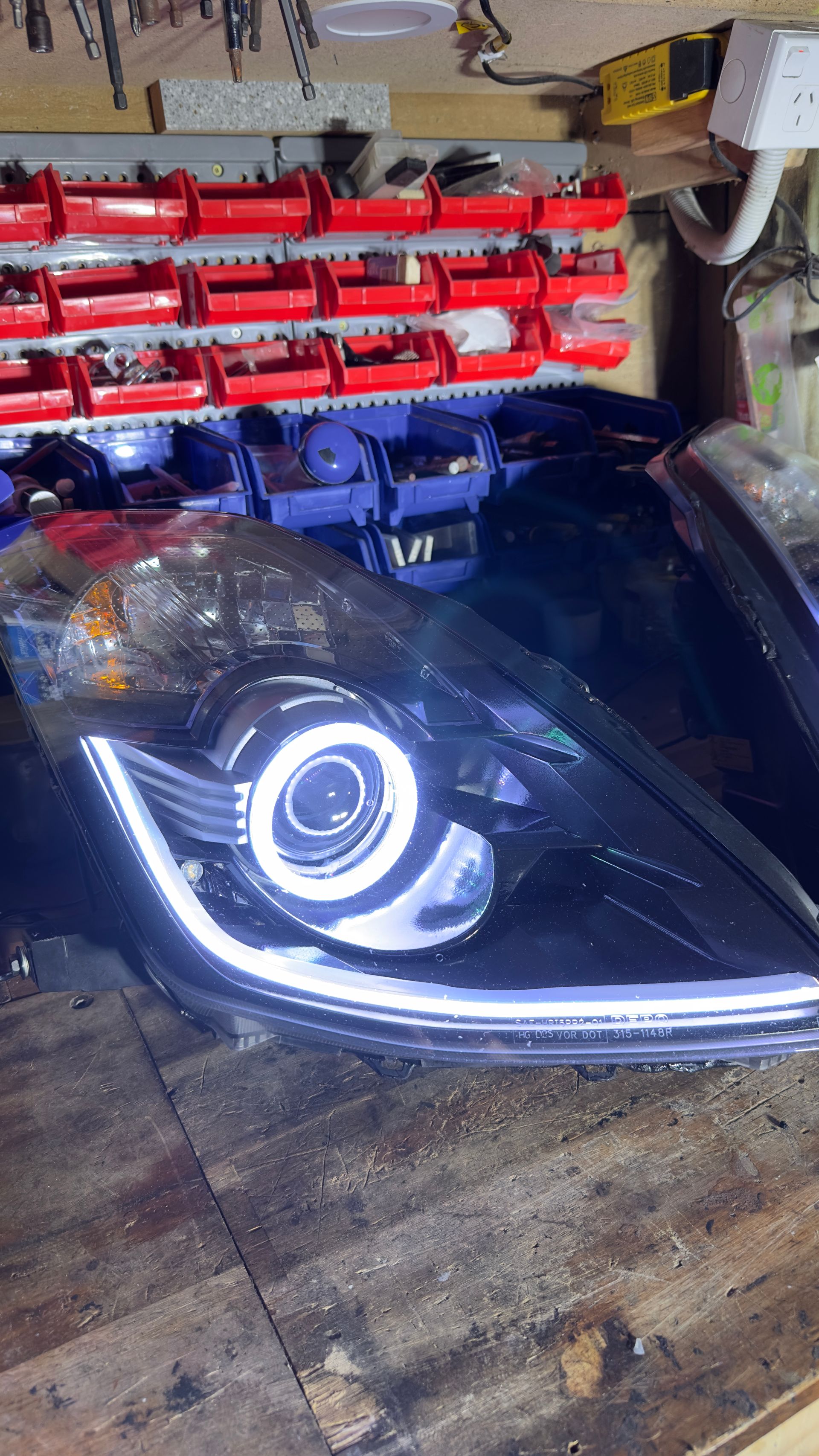 Black car headlight with a white halo and LED strip on a wooden surface in a workshop.