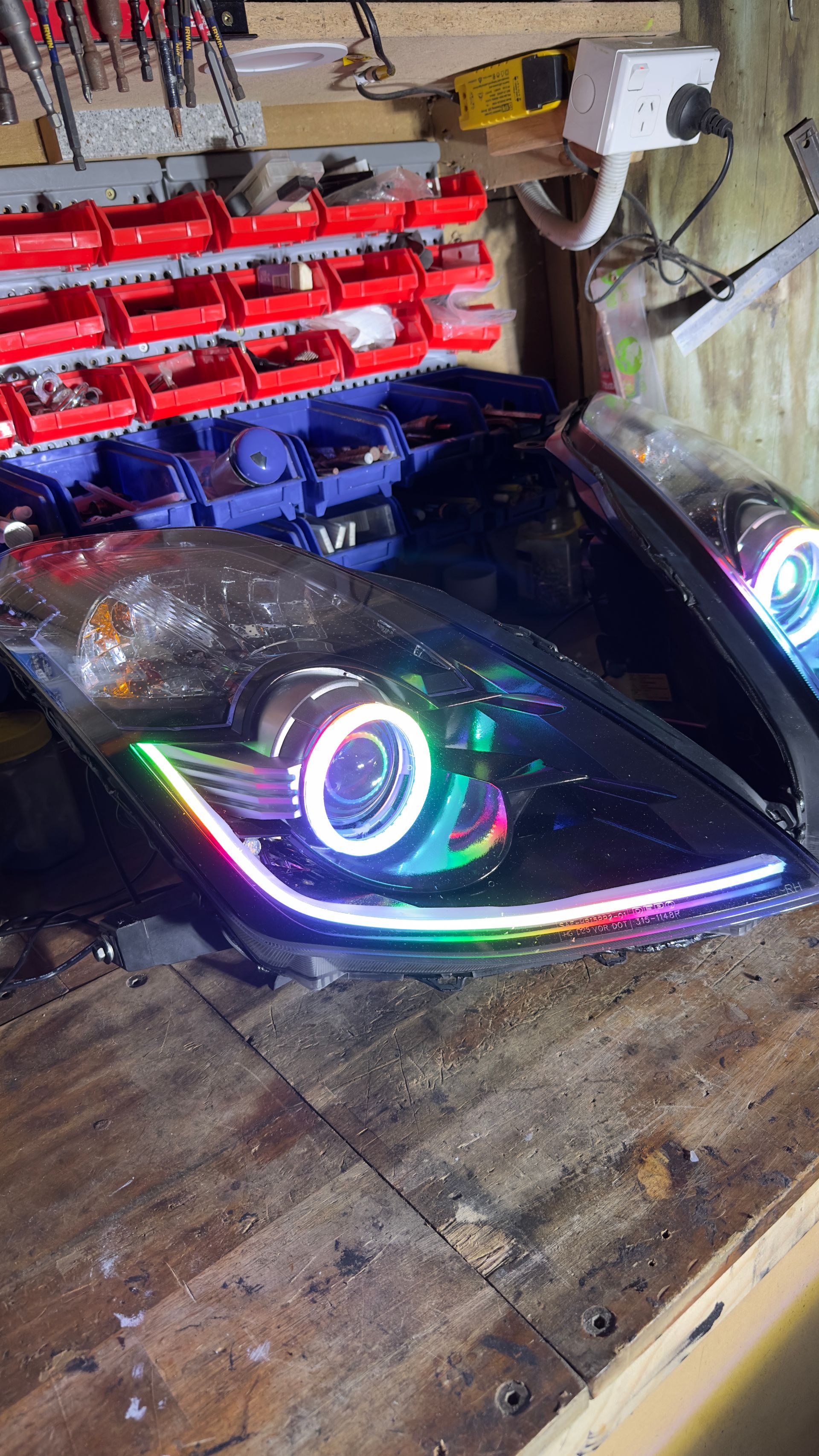 Black car headlight with a white halo and LED strip on a wooden surface in a workshop.