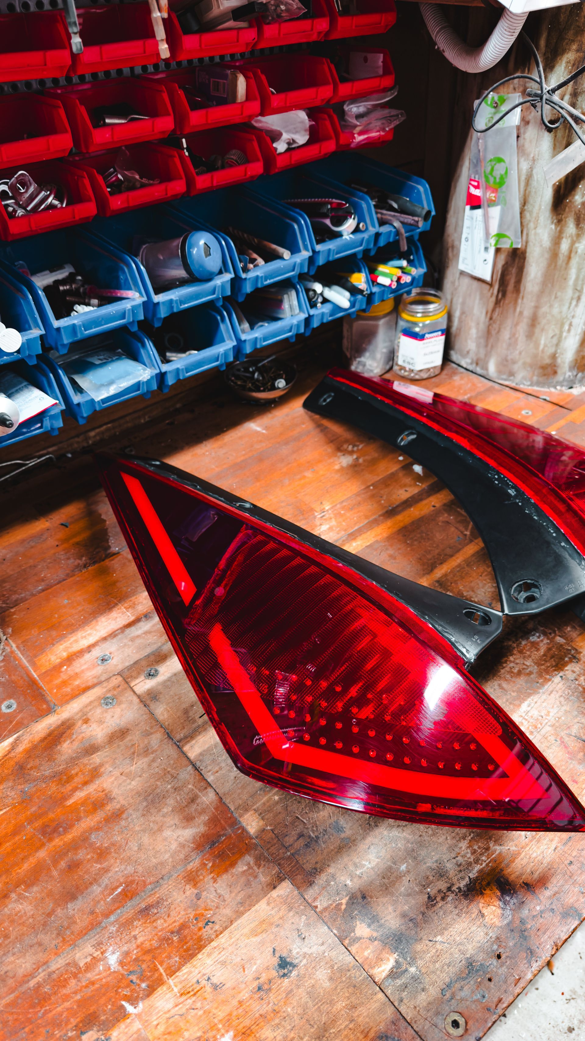 Red car taillight components on a wooden surface, with a parts storage system in the background.