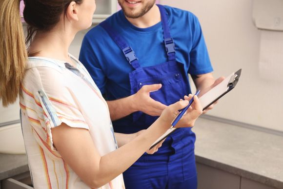 A man in blue overalls is talking to a woman in a kitchen while holding a clipboard.