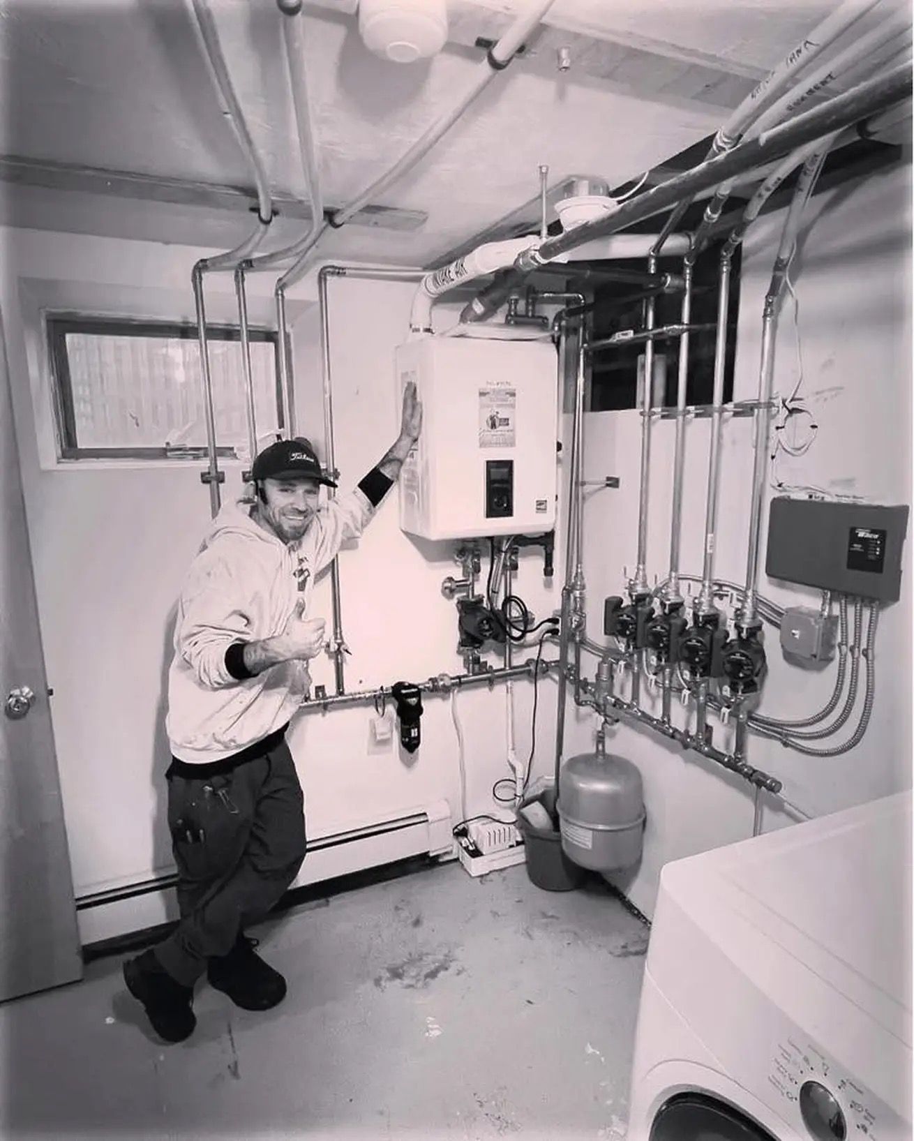 Man standing next to a water heater in a utility room, pointing up. There are pipes and a washing machine.
