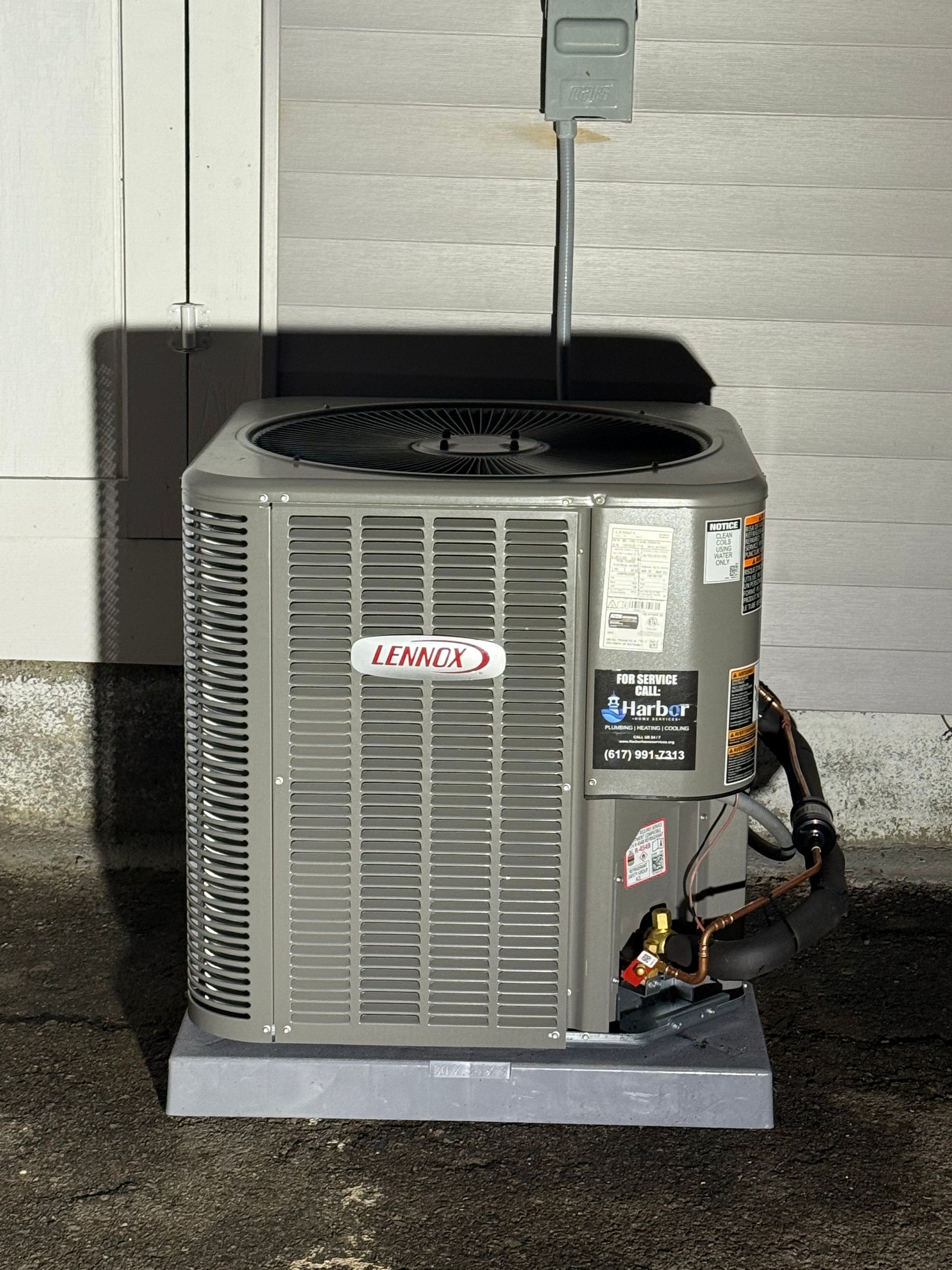 Lennox air conditioning unit on a concrete pad next to a light-colored wall, with electrical box above.