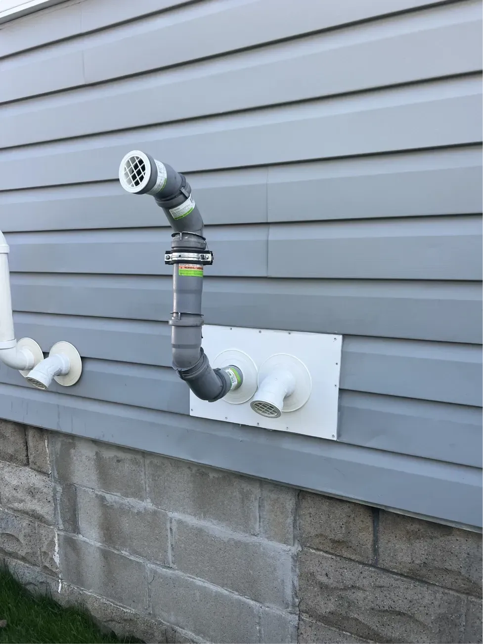 Exterior view of a gray pipe venting from a white rectangular opening on light gray siding wall above a concrete foundation.