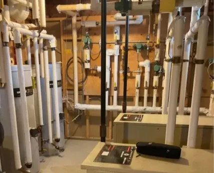 Heating system with white pipes, beige boxes, and black pipes in a utility room.