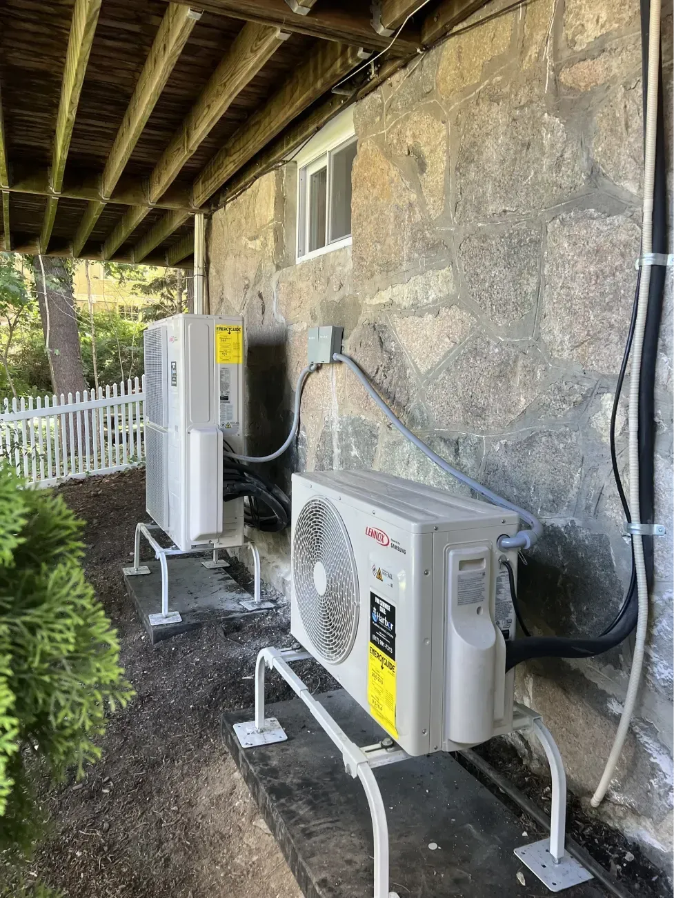 Two white HVAC units mounted on a concrete wall under a covered porch.
