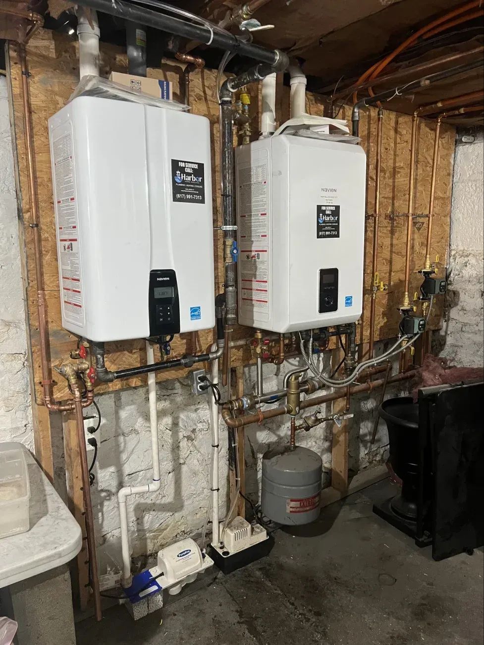 Two white tankless water heaters mounted on a wooden wall, with copper pipes and a gray expansion tank.