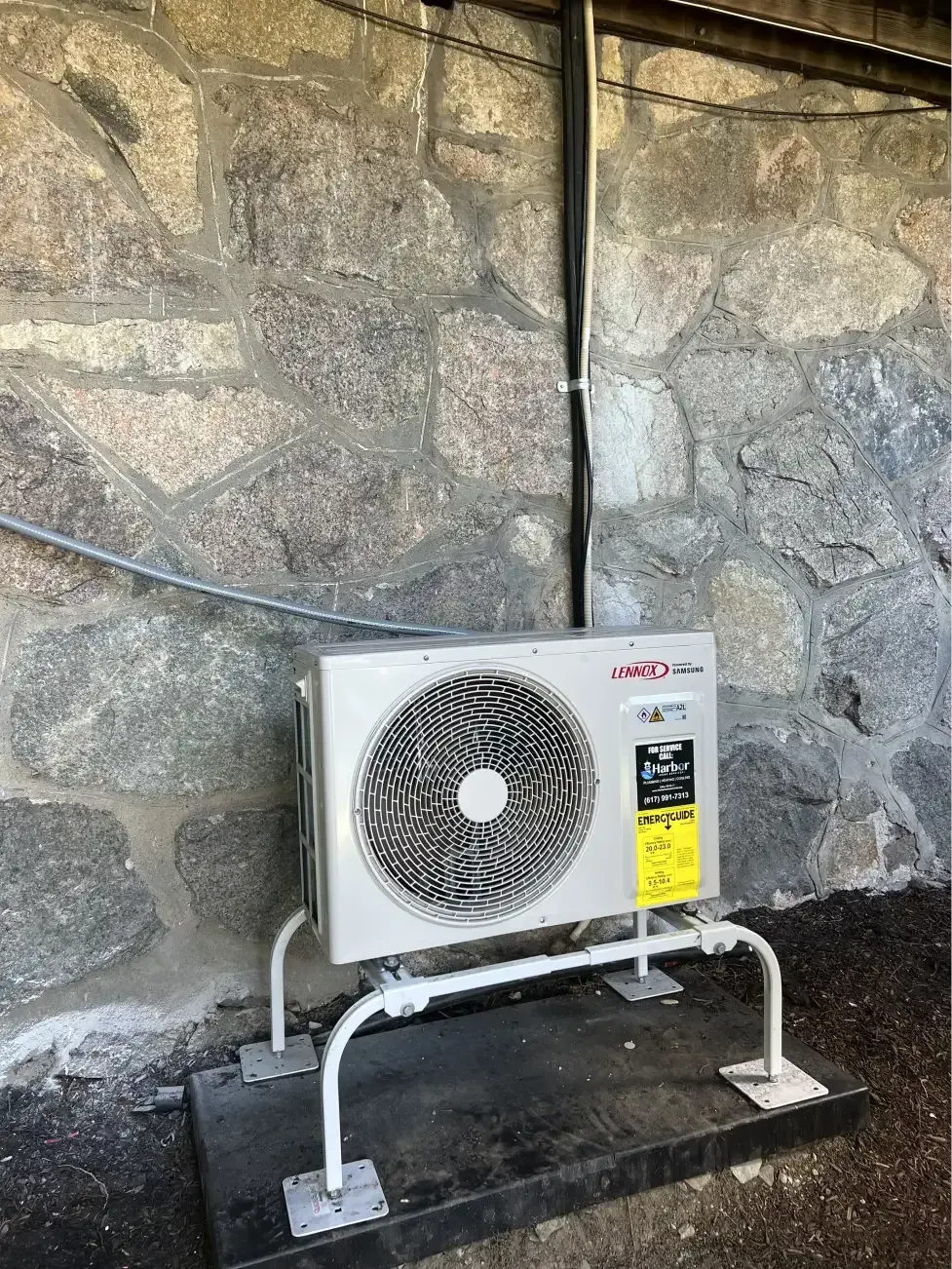 An outdoor air conditioning unit on a stone wall. It's mounted on a metal stand and has a black conduit running above.