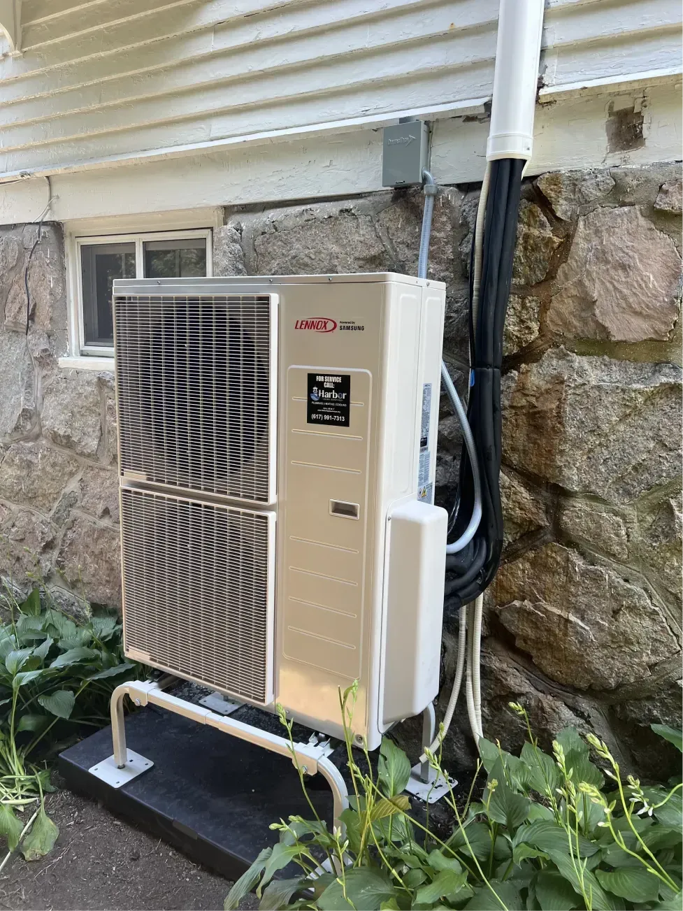 A beige and black outdoor heat pump unit attached to a stone wall with greenery around it.
