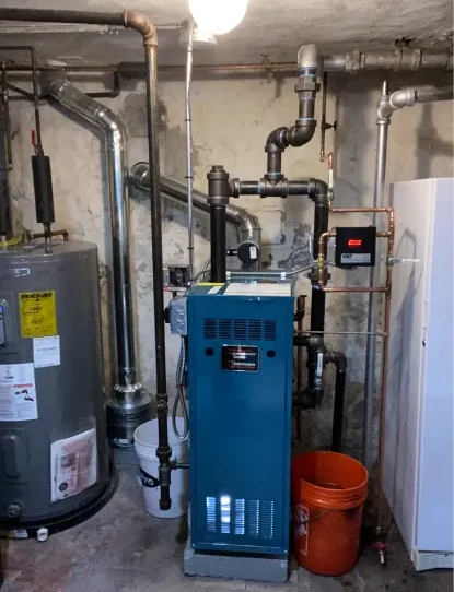 A basement with a blue furnace, hot water tank, pipes, and orange bucket. Concrete walls.