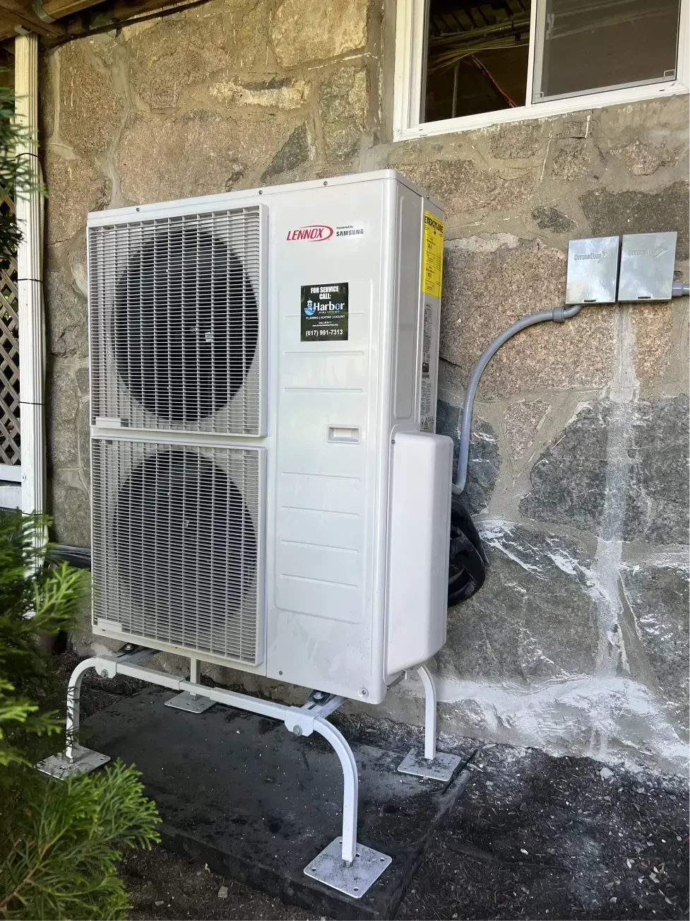 Air source heat pump on exterior of building. White unit, mounted on a metal frame against stone wall.