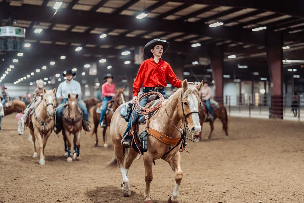 Boy on horse in arena, wearing cowboy hat, blue shirt, jeans. Horse is dark with white face.