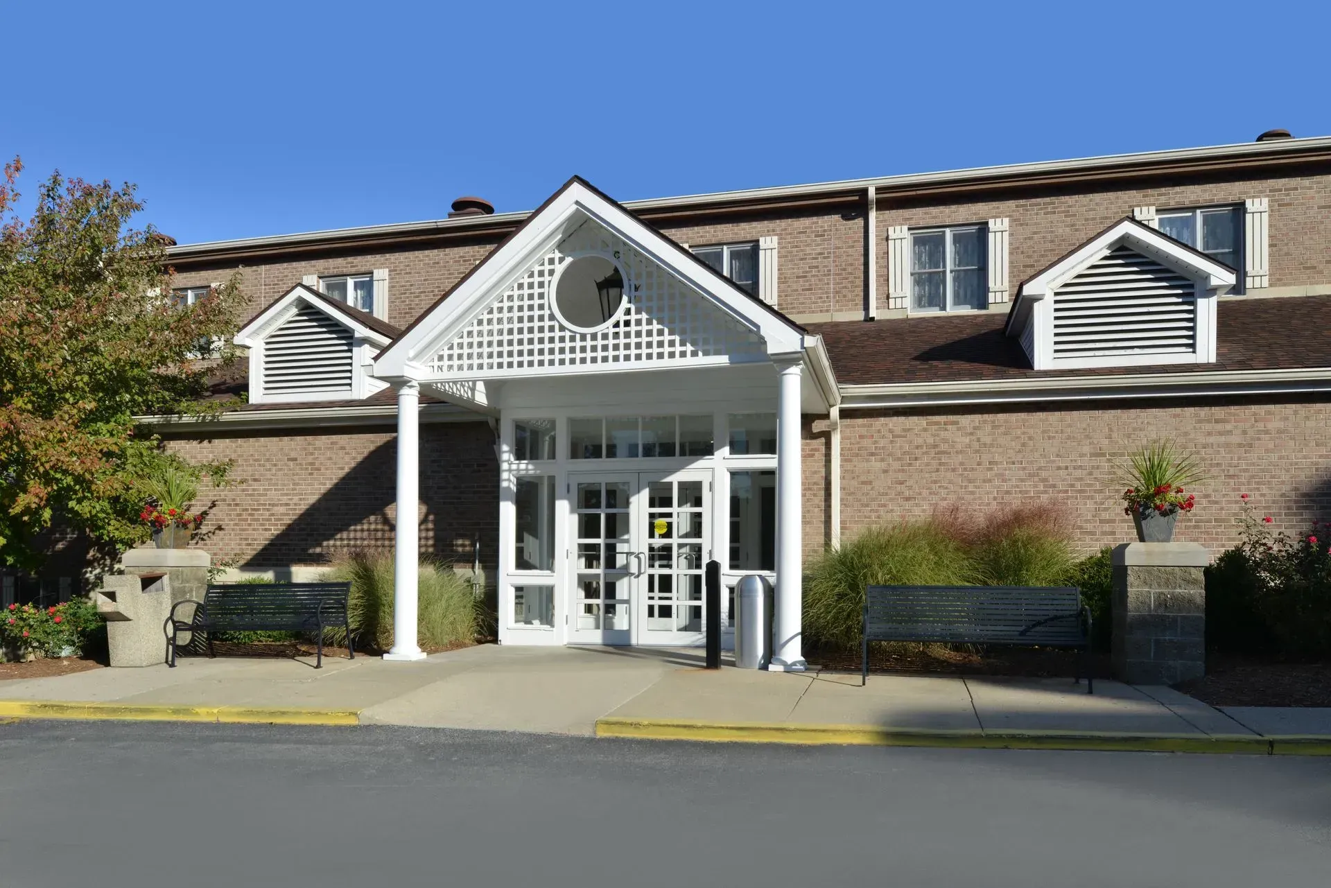 Brick building with a white columned entrance, glass doors, and two benches on a concrete sidewalk under a clear sky.