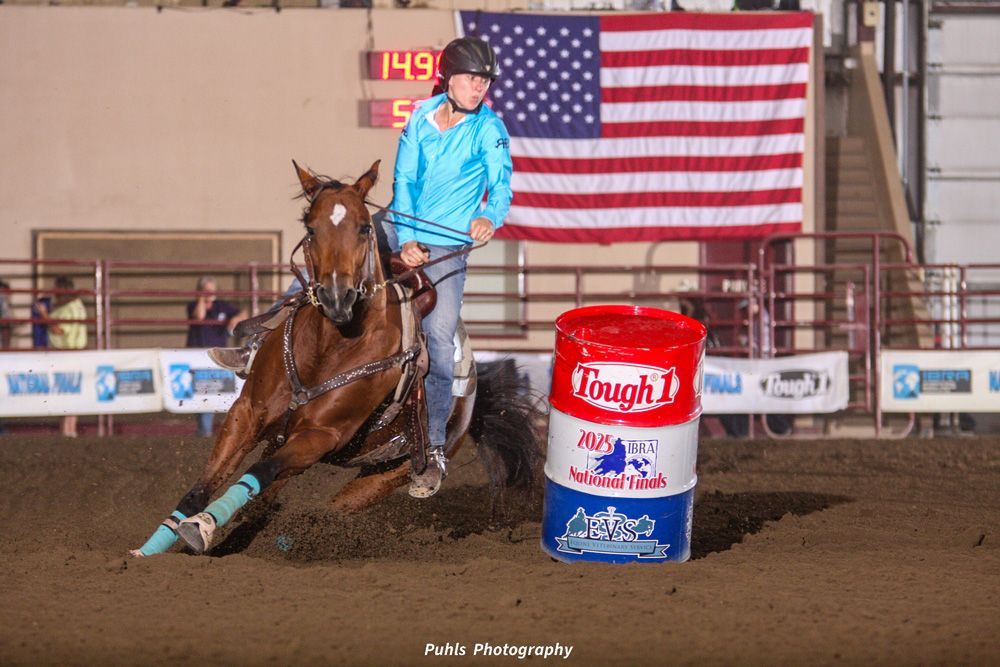 Horse and rider rounding a barrel in an indoor rodeo arena with a U.S. flag backdrop