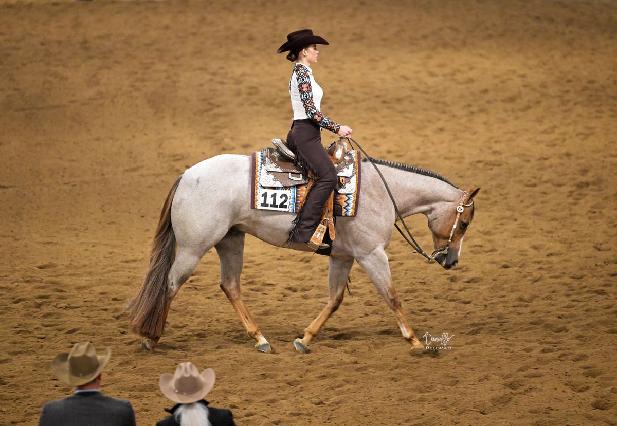 A person wearing a cowboy hat and western show clothes rides a light-colored horse across an arena dirt floor.