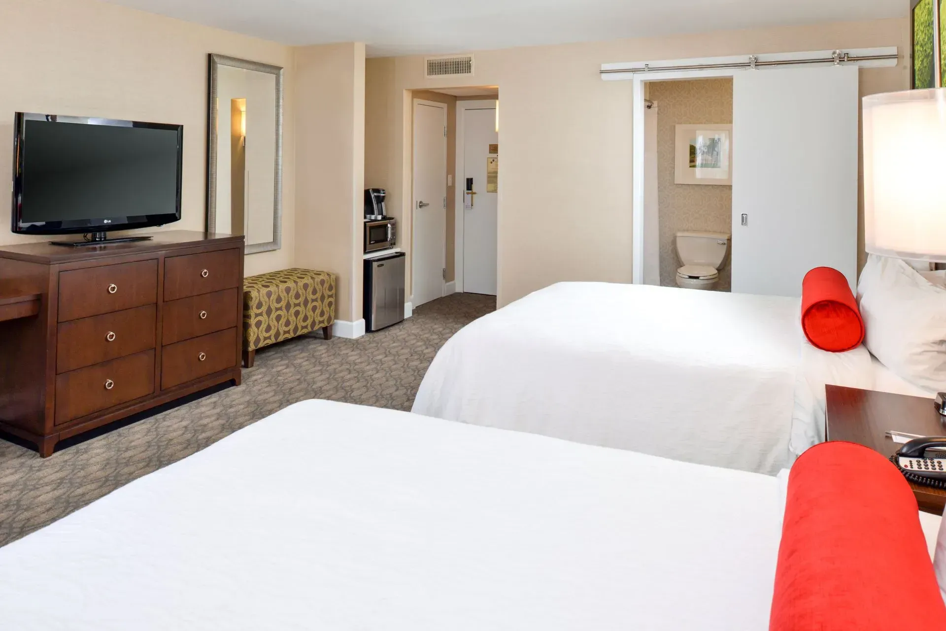 A hotel room with two white beds, a wooden dresser, a television, and a sliding door revealing a bathroom.
